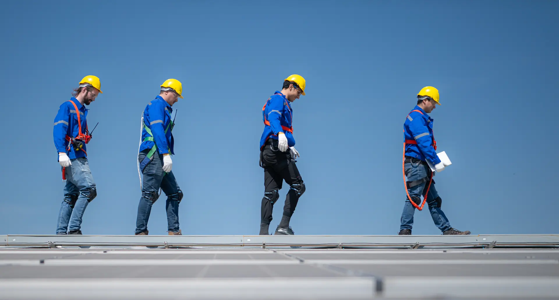 Four solar panel installers in hard hats walking across a rooftop array against a clear blue sky